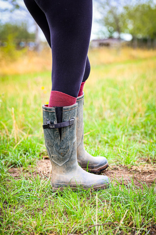 A close-up outdoor shot of a person wearing tall gray rubber rain boots with a black strap and buckle, paired with dark leggings and Soleberie Burgundy Classic Long Socks, standing in a grassy field.