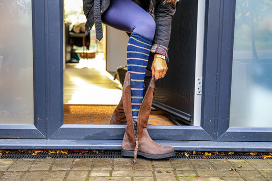 A person wearing Navy Stripe Classic Long Socks from Soleberie and brown suede knee-high boots and a dark jacket, standing in front of a glass door.