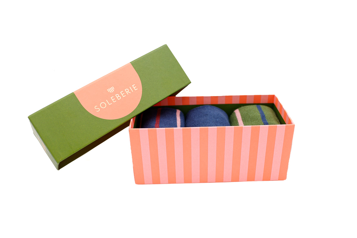 An open Soleberie gift box with a green lid and pink and coral striped sides, containing three pairs of Soleberie Classic Long Socks in navy, green and navy stripe. The lid has a pink label reading Soleberie.