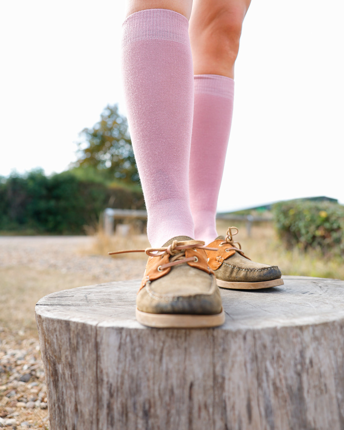 A person standing on a tree stump outdoors, wearing Soleberie Pink Classic Long Socks and tan boat shoes.